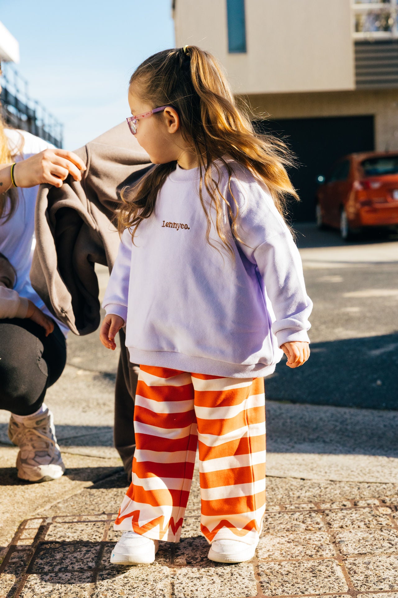 Child wearing a lilac purple sweater and red striped pants standing on a sidewalk for a photoshoot