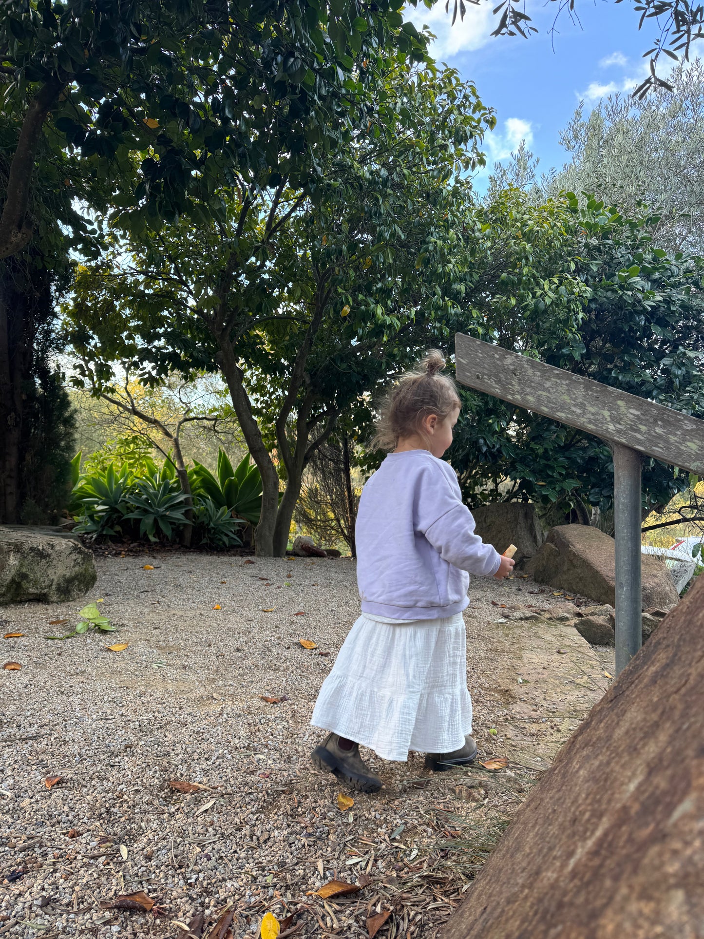 Child in a white dress standing outdoors near trees and rocks, little girl wearing white skirt and purple jumper