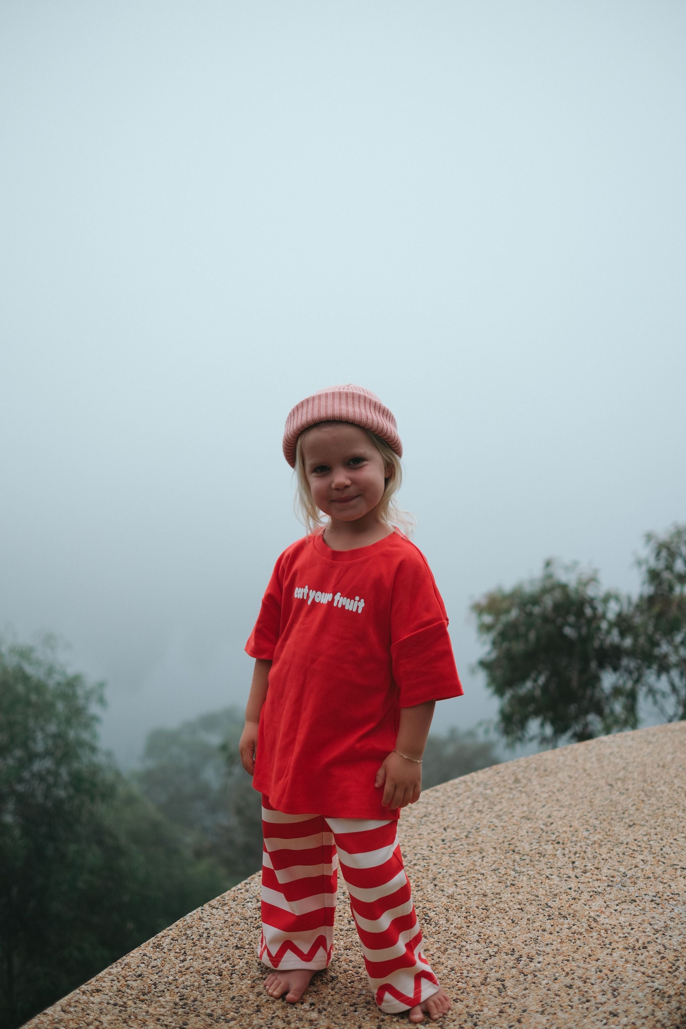child standing on cliff wearing red 'eat your fruit' tee and red striped pants with lightning strike