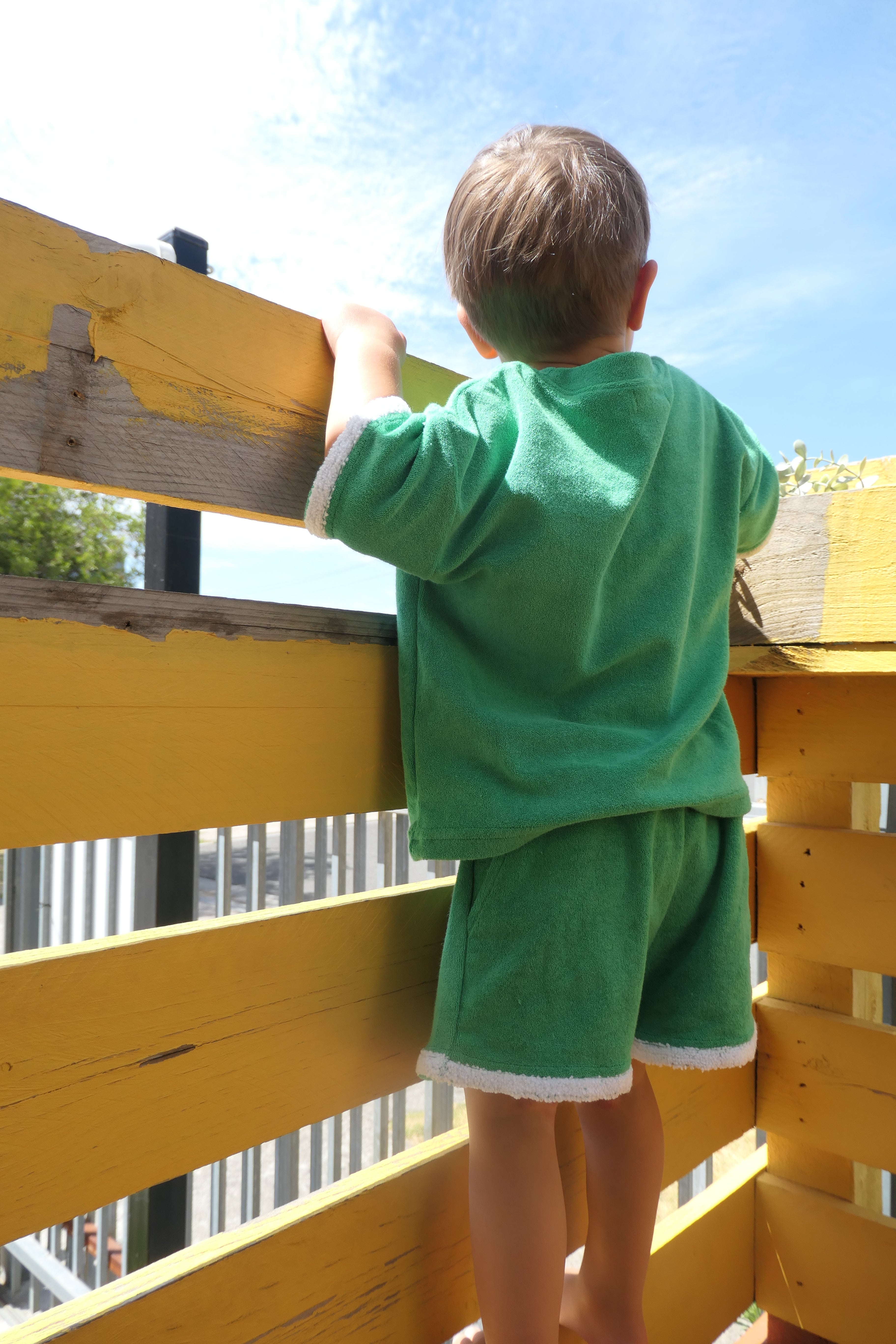 Child in a green terry towellong outfit standing on a yellow wooden outdoor play equipment with a blue sky background
