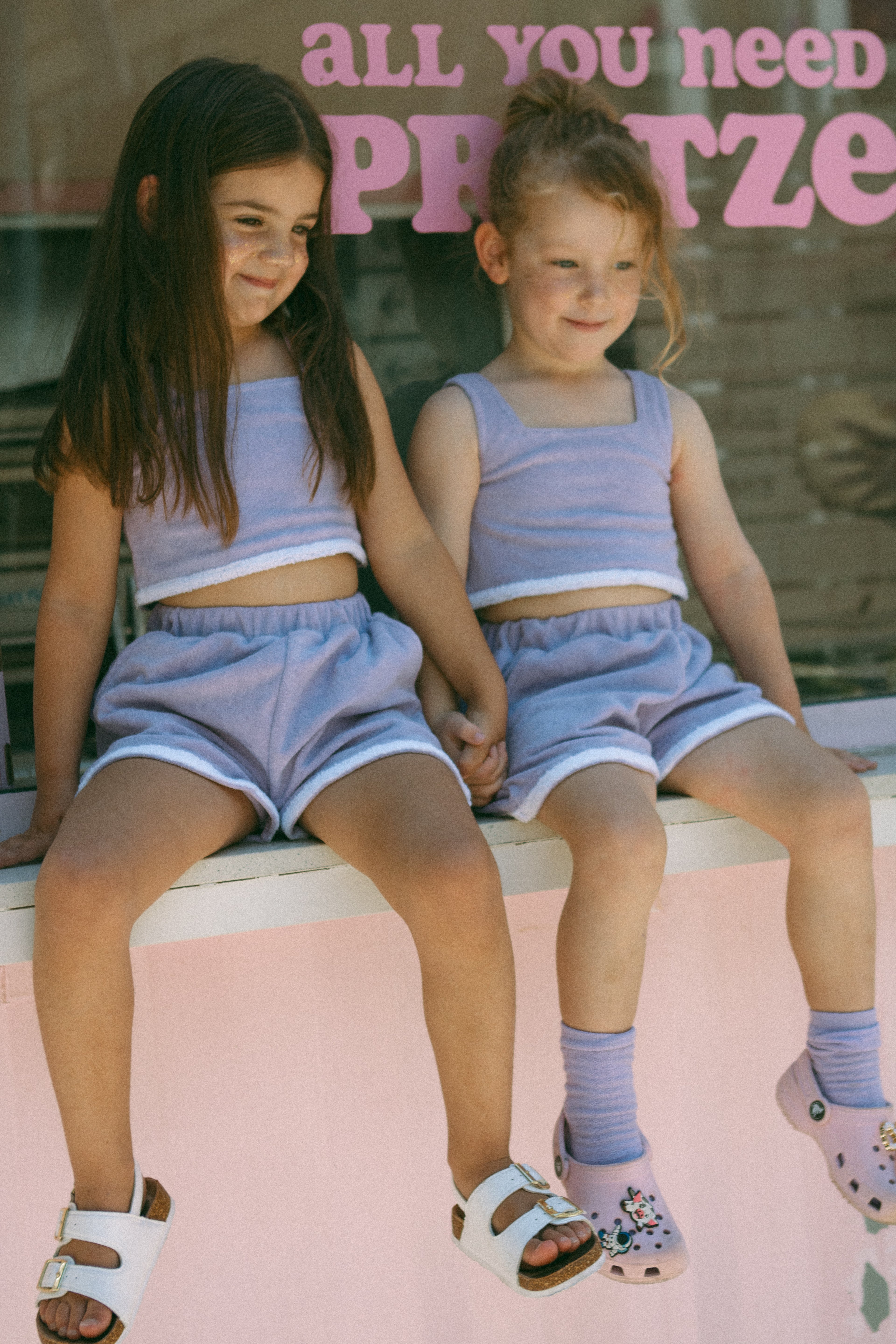 Two young girls in matching lilac outfits sitting on a ledge with text in the background.