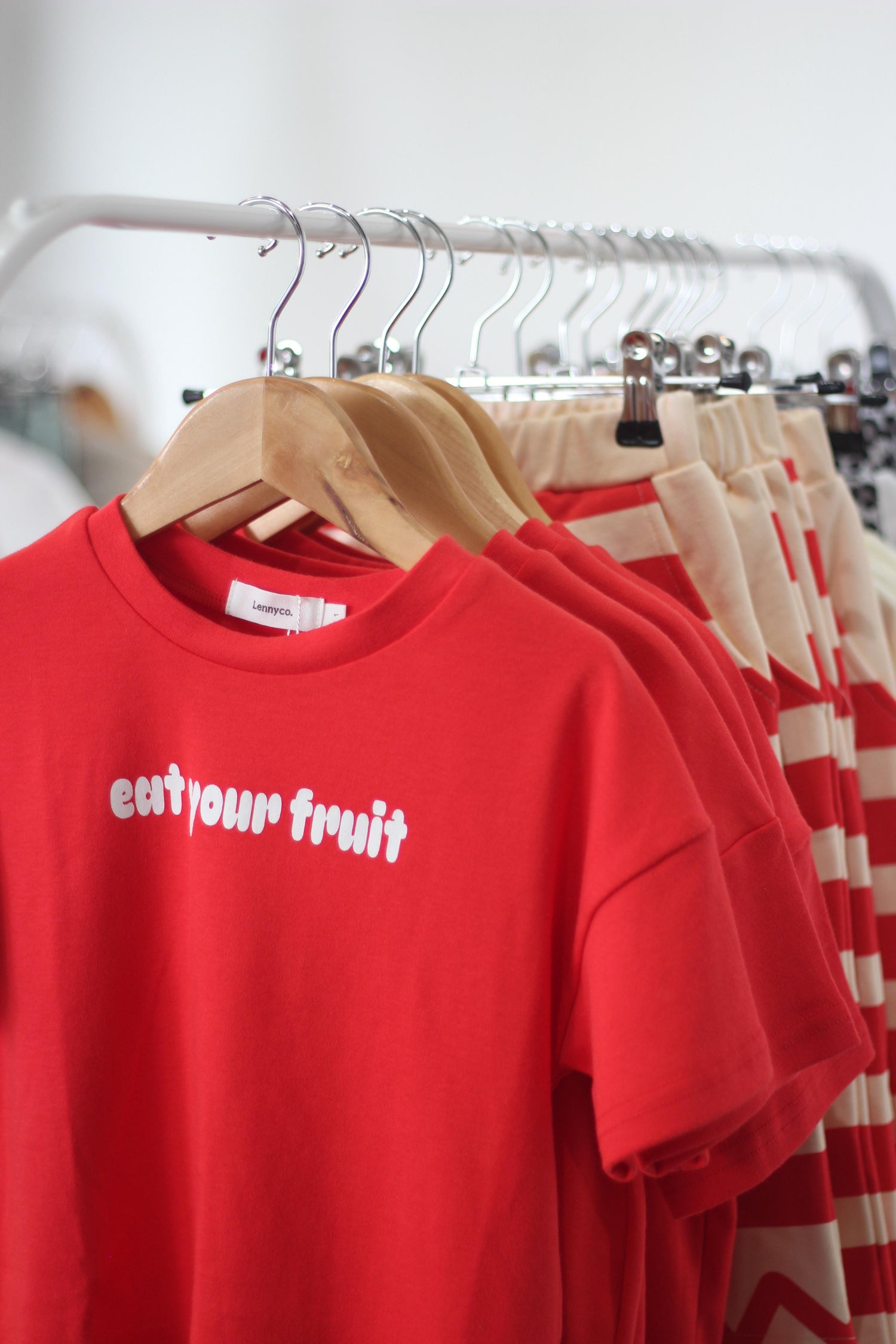 A red t-shirt with the text 'EAT YOUR FRUIT' printed on the front, displayed among other clothing items, kids fruit tee